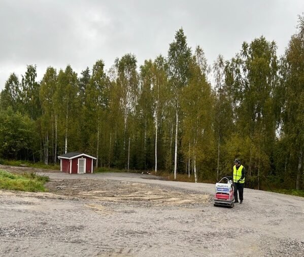 En person i gul v&auml;st st&aring;r vid en maskin p&aring; en grusv&auml;g, med en r&ouml;d stuga och bj&ouml;rkskog i bakgrunden.