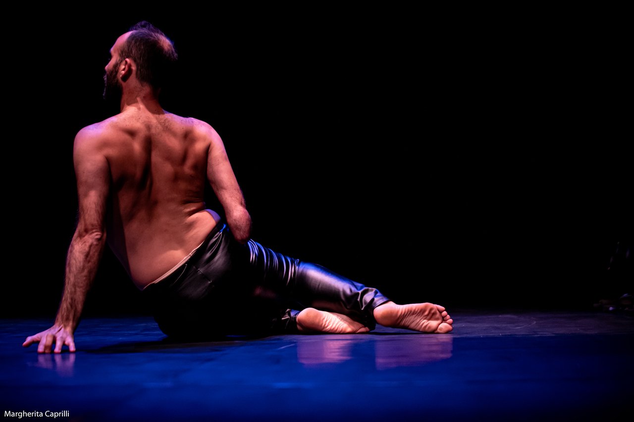 Dansare sitter på golvet med ryggen mot kameran, i sidolutad position under mörk scenbelysning