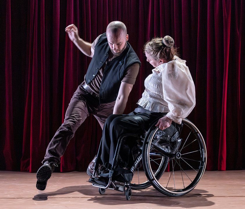 Två dansare uppträder på scen framför röda ridåer; en sitter i rullstol och den andra står, båda i en intensiv rörelse.