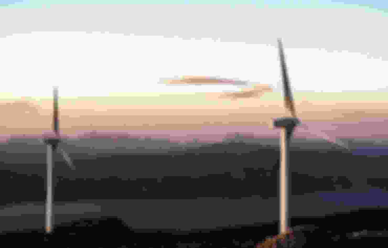 Two wind turbines stand out in front of a beautiful background of mountains during sunset.