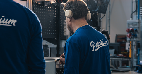 A man with his back turned to the camera is wearing a shirt with the Polarium-logo on the back. He’s wearing over ear-headphones that aren’t properly on because he’s pointing to something whilst talking to a coworker who is standing beside him.