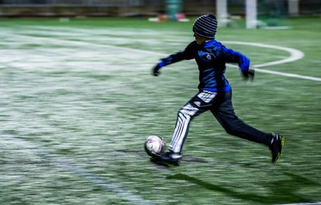 Pojke i mössa och vantar springer med en fotboll i i svagt elljussken.