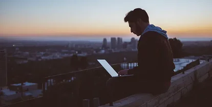 En man sitter på ett hustak i en storstad vid soluppgången och tittar på sin laptop.