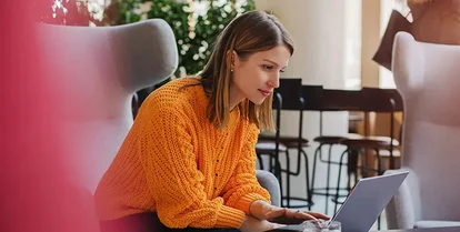 En kvinna i orange tröja sitter på ett kontor med sin laptop.