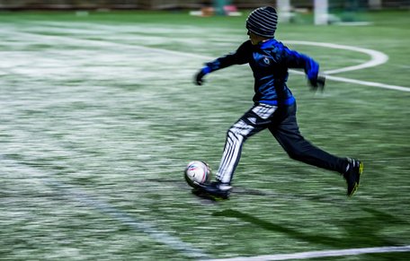 Pojke i mössa och vantar springer med en fotboll i i svagt elljussken.