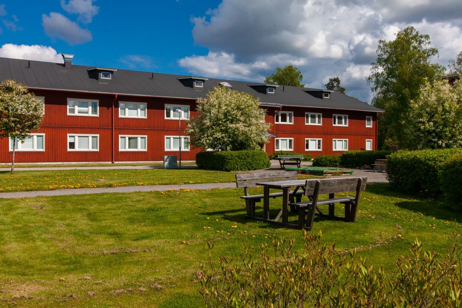 Exterior of red, twostorey buildings with green grass lawn infront. A table and benches on the lawn and trees and bushes around.