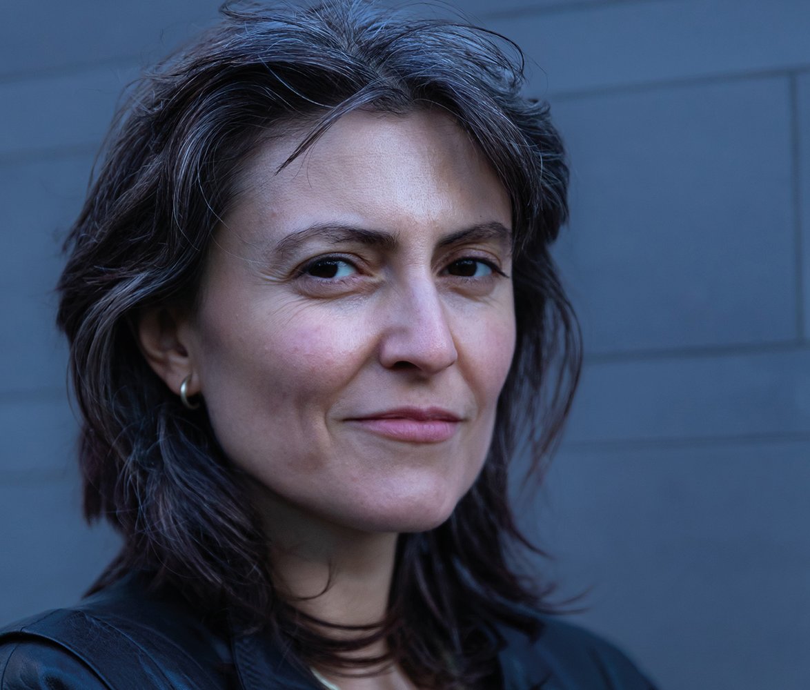 Porträtt av en kvinna med kort mörkt hår och läderjacka som står mot en neutral, grå vägg och ser lugnt in i kameran.