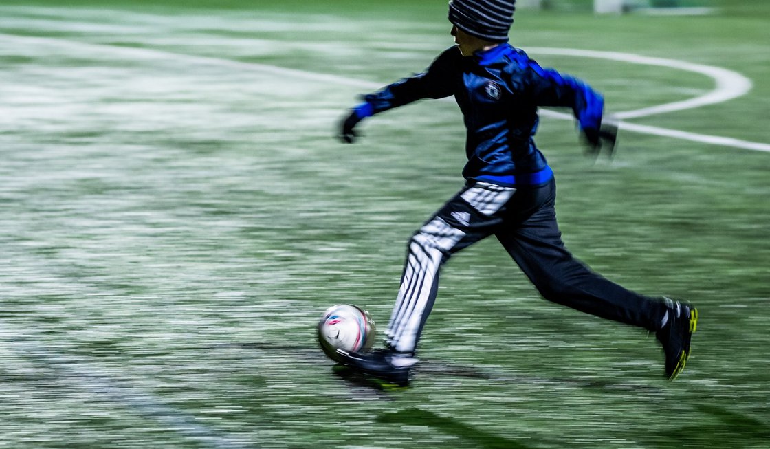 Pojke i mössa och vantar springer med en fotboll i i svagt elljussken.