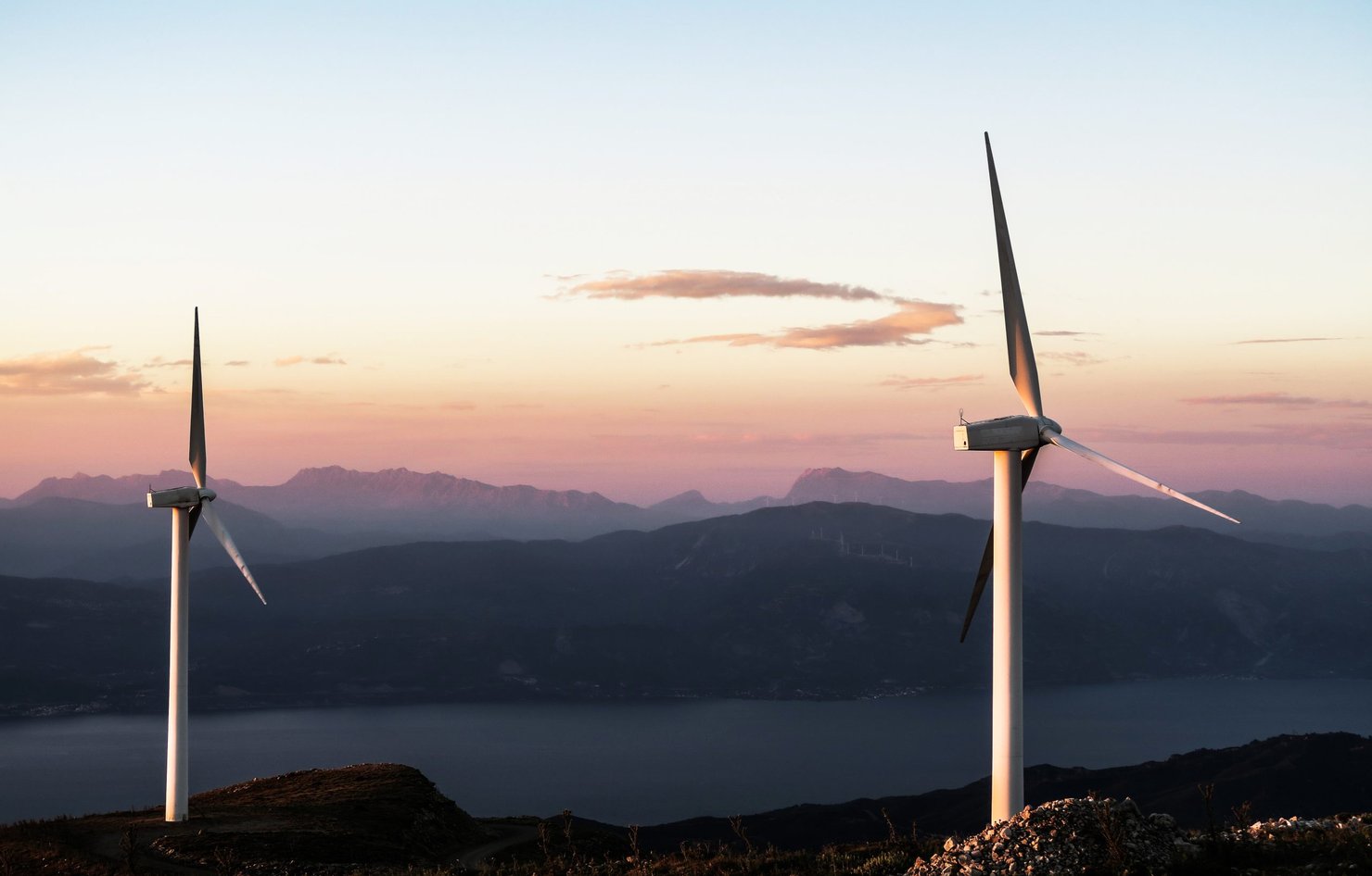 Two wind turbines stand out in front of a beautiful background of mountains during sunset.