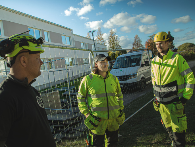 Tre arbetare i gul reflektvast diskuterar vid byggplats med fordon och byggnad i bakgrunden.