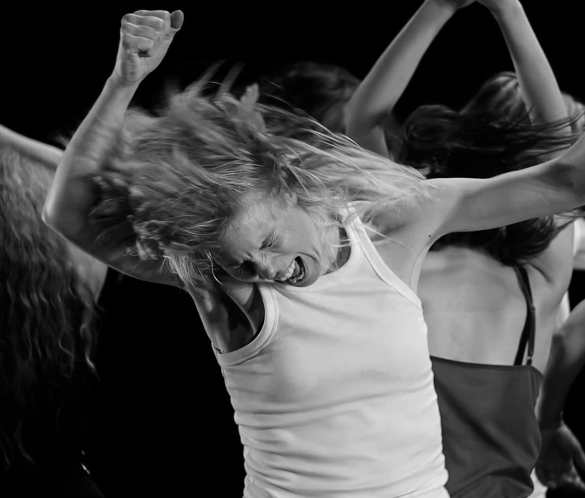 Kvinna med vildsint uttryck dansar med knutna nävar i luften, fler dansare syns i bakgrunden.