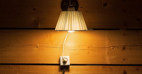 A lit lamp hanging on a wooden wall, plugged in slightly underneath where it’s hanging. The lampshade is white and crinkly, with a farmhouse feel to it.