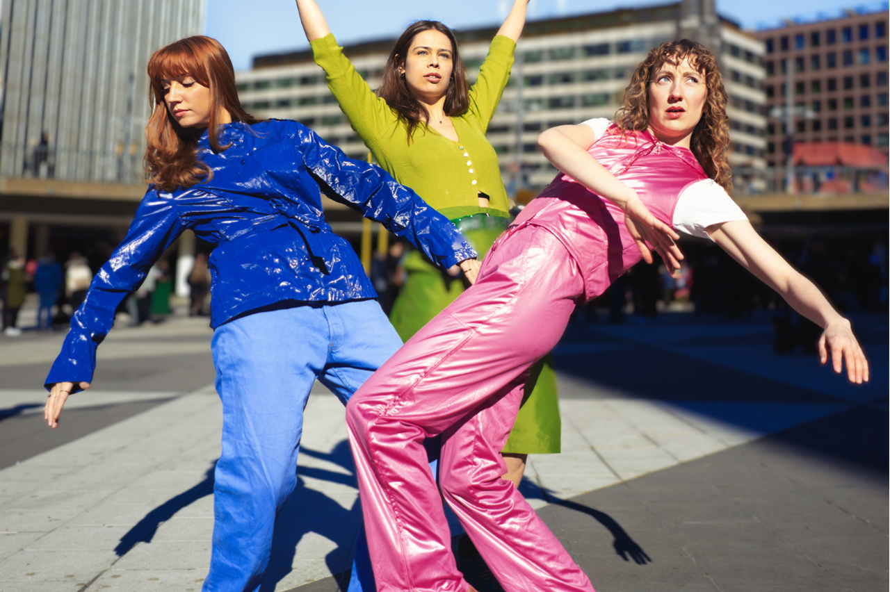 Bild på gruppen Dansbandet som dansar på Sergels Torg med stora byggnader i bakgrunden.