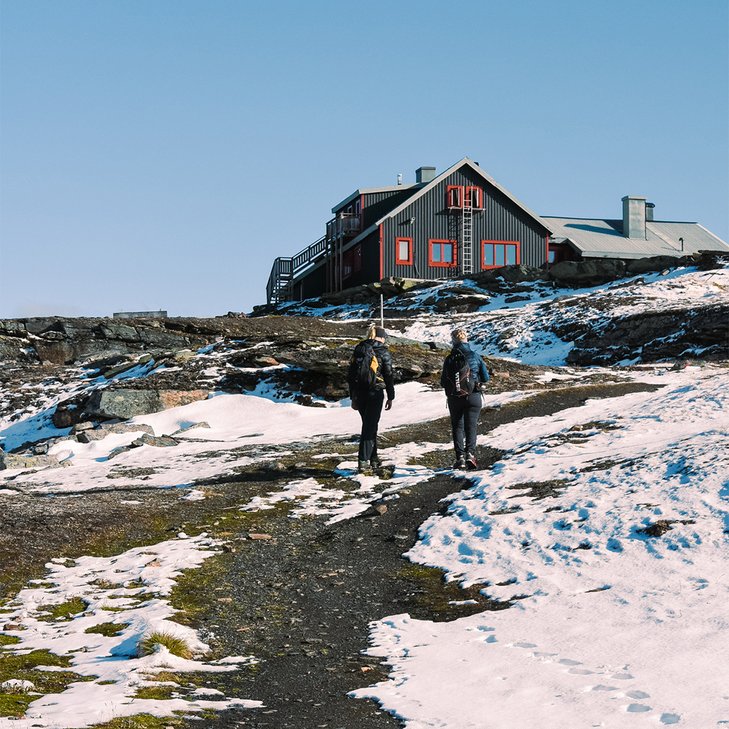 Two women are walking the final stretch along a snow-flecked trail up to the Låktatjåkko Mountain Station