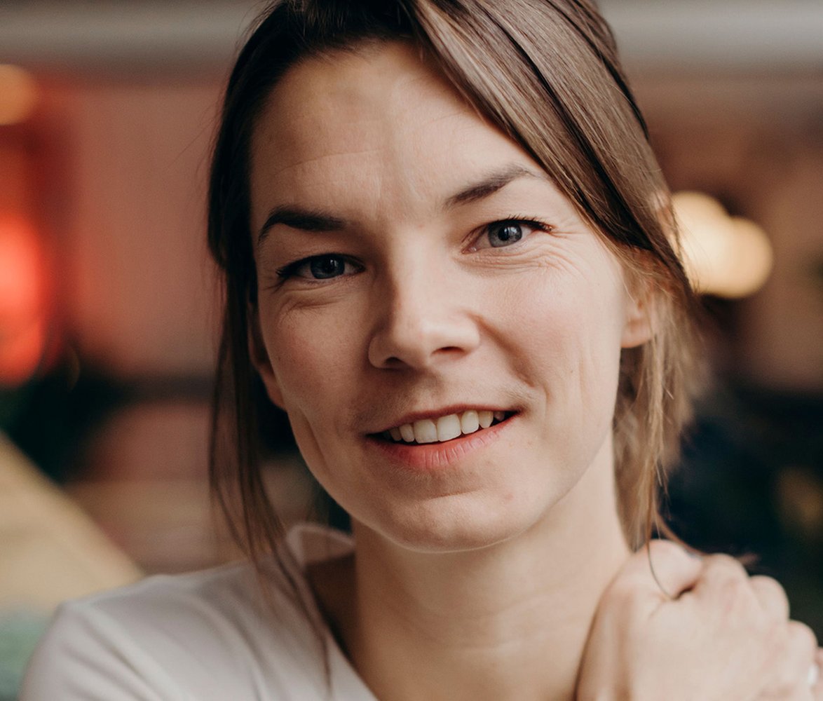 Porträttbild av en leende kvinna med ljust hår som tittar in i kameran. Bakgrunden är mjukt suddig med varma färgtoner.