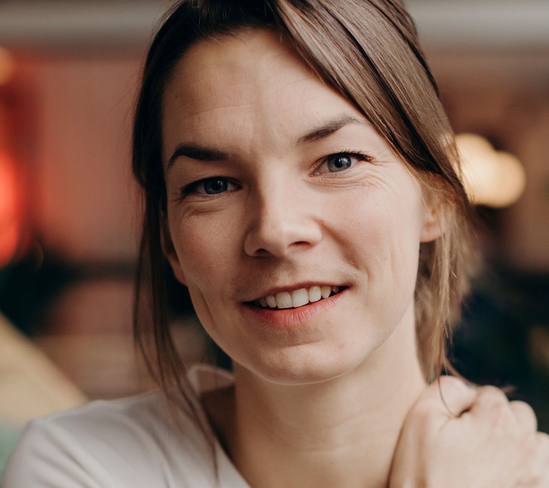 Porträttbild av en leende kvinna med ljust hår som tittar in i kameran. Bakgrunden är mjukt suddig med varma färgtoner.