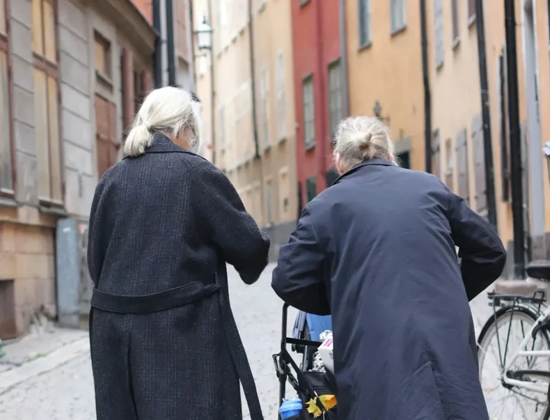 Två äldre kvinnor går i gamla stan med en barnvagn.