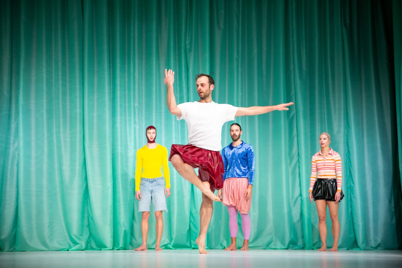 Dansare i mitten balanserar på ett ben med armarna utsträckta, medan fyra andra står bakom i stillhet.