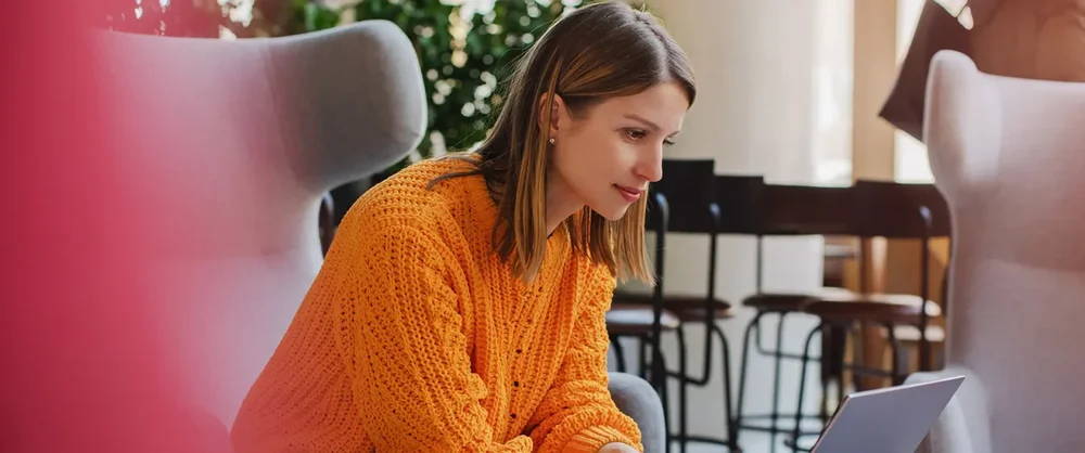En kvinna i orange tröja sitter på ett kontor med sin laptop.