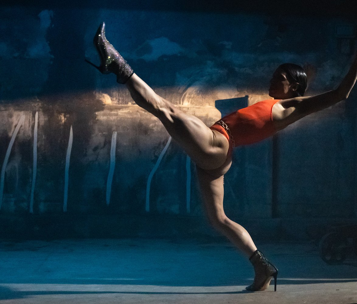 Dansaren Daniel Proietto står i röd dräkt och glittriga högklackade skor balanserar i en dramatisk danspose i ett dunkelt, industriellt rum med blått ljus.