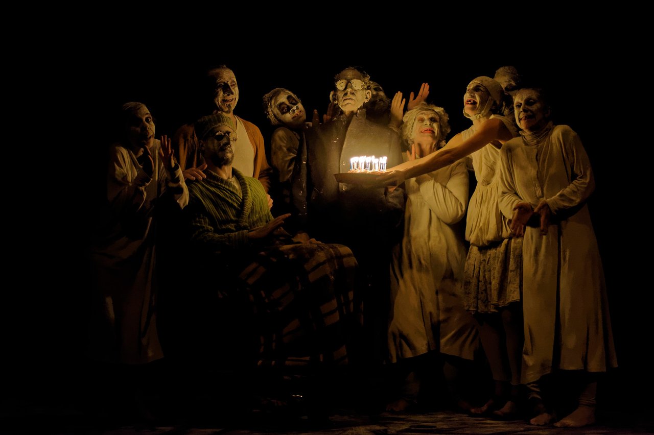 En grupp människor med vitsminkade ansikten, som räcker fram en födelsedagstårta mot en person.