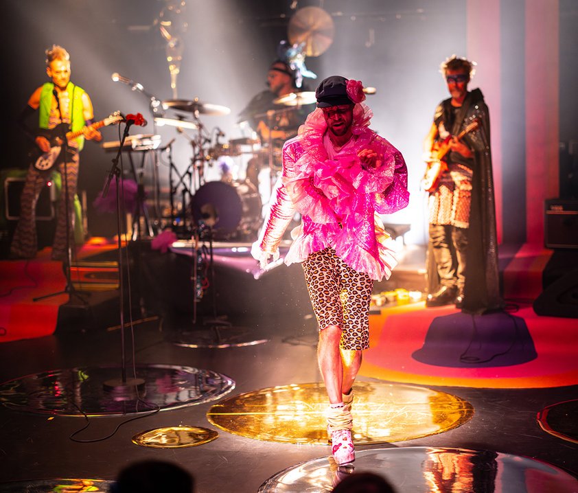 Ett rockband på scen och en person i mitten med rosa glittrig blus.