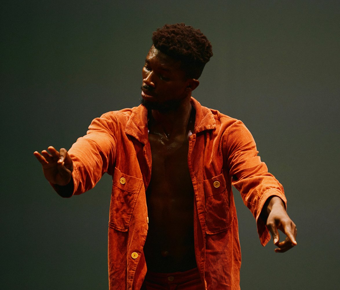 En man med mörk hy i orange kläder står ensam på en scen med utsträckta armar i en mjuk, kontrollerad rörelse mot mörk bakgrund.