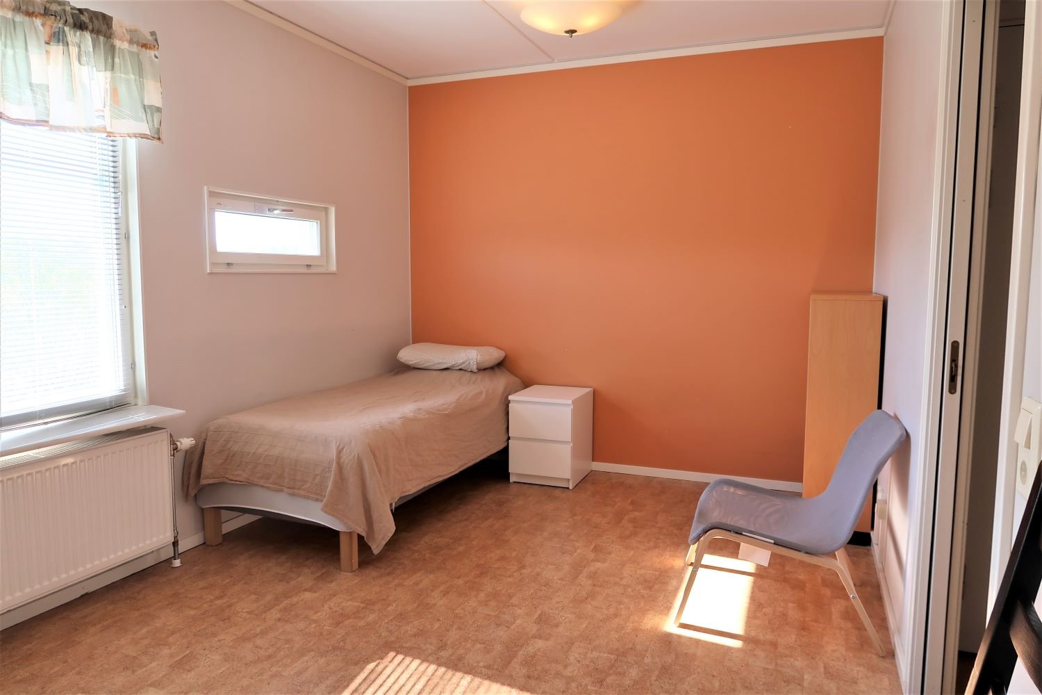 studentroom with orange wall, bed, chair, bookshelf. and bedside table.