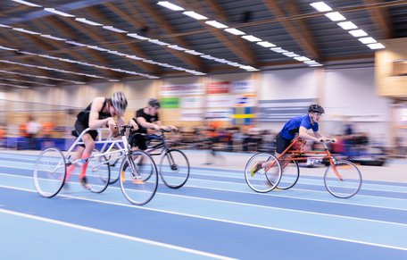 Tre deltagare tävlar i framerunning inomhus på en löparbana. Cykeln har ett hjul fram och två bak.