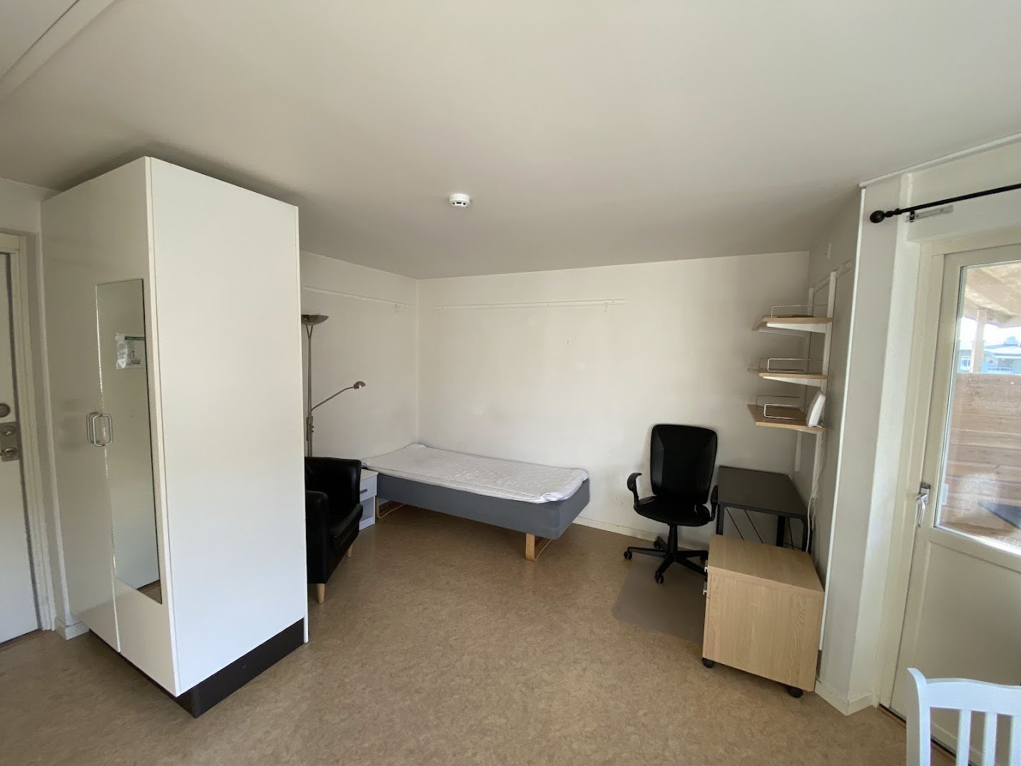 Student room with wardrobes, bed, desk, desk chair, chest of drawers and door to balcony.