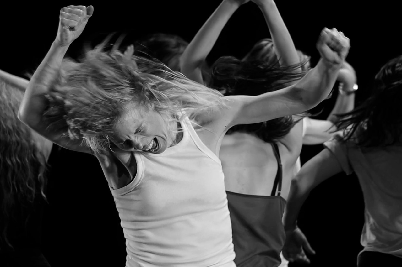 Kvinna med vildsint uttryck dansar med knutna nävar i luften, fler dansare syns i bakgrunden.