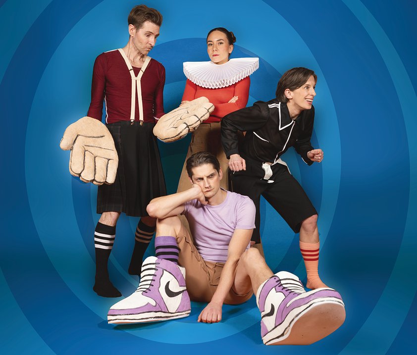 Fyra personer i lekfulla, stiliserade kostymer poserar tillsammans mot blå bakgrund, med överdimensionerade accessoarer och uttrycksfulla poser.