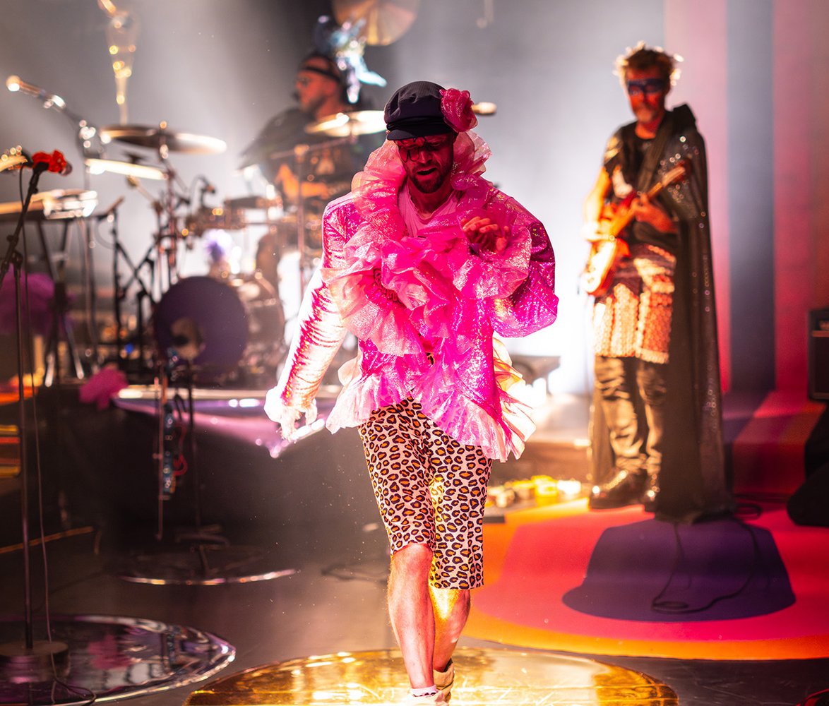 Ett rockband på scen och en person i mitten med rosa glittrig blus.