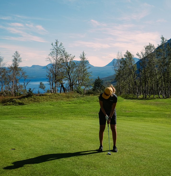 kvinna måttar med sin putter på golfbanan en solig dag med Lapporten i bakgrunden