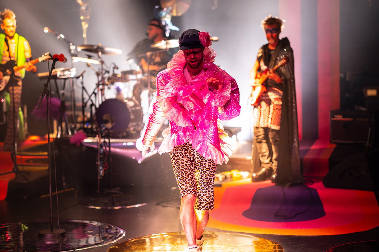 Ett rockband på scen och en person i mitten med rosa glittrig blus.