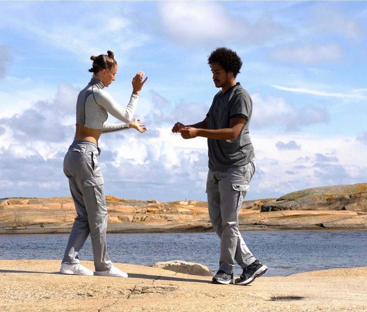 En kvinna med uppsatt hår i form av tre knutar på huvudet samt en mörkhyad man där båda har gråa kläder utför långsamma, koncentrerade rörelser mot varandra på klippor vid havet under en ljus himmel.