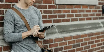 En man i grå tröja står lutad mot en tegelvägg och skriver på sin surfplatta.