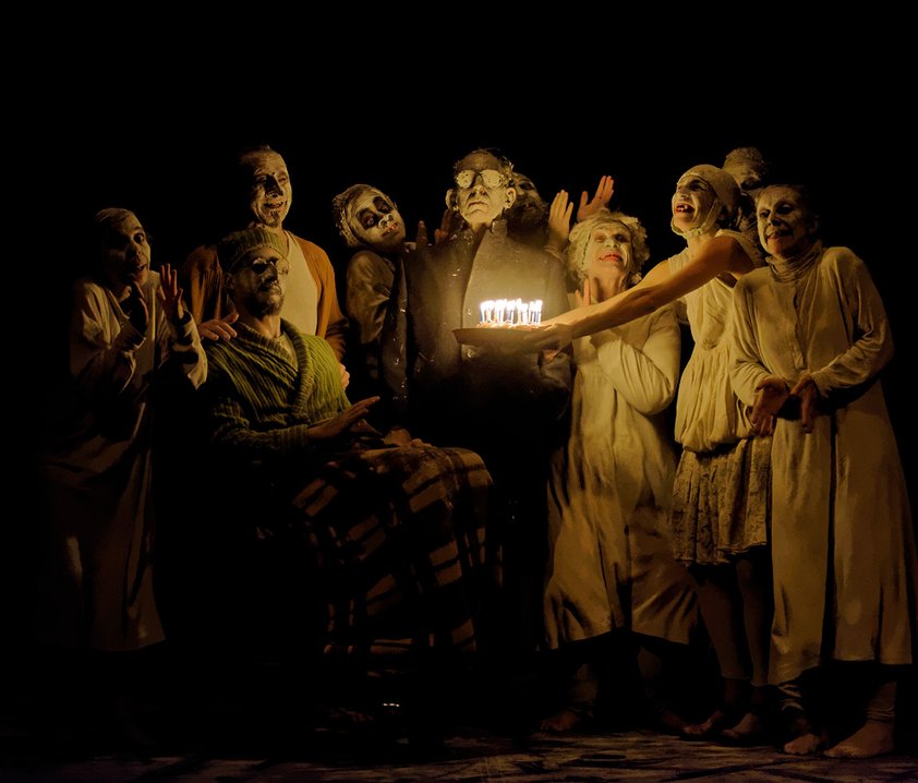 En grupp människor med vitsminkade ansikten, som räcker fram en födelsedagstårta mot en person.