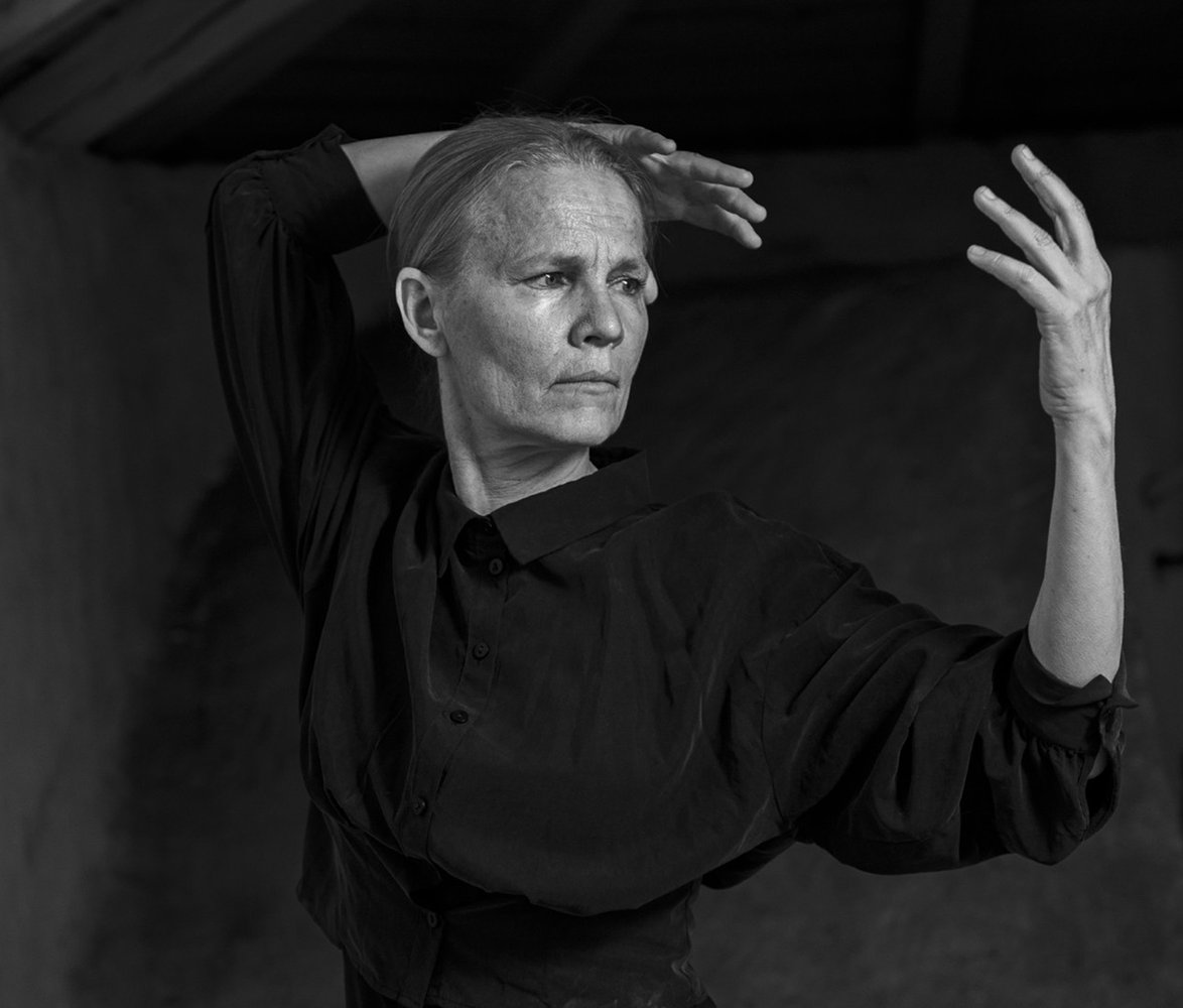 Svartvit bild av en kvinna med håret kammat bakåt, hon har en långärmad skjorta som håller armarna lyfta i en kontrollerad, uttrycksfull danspose inomhus.