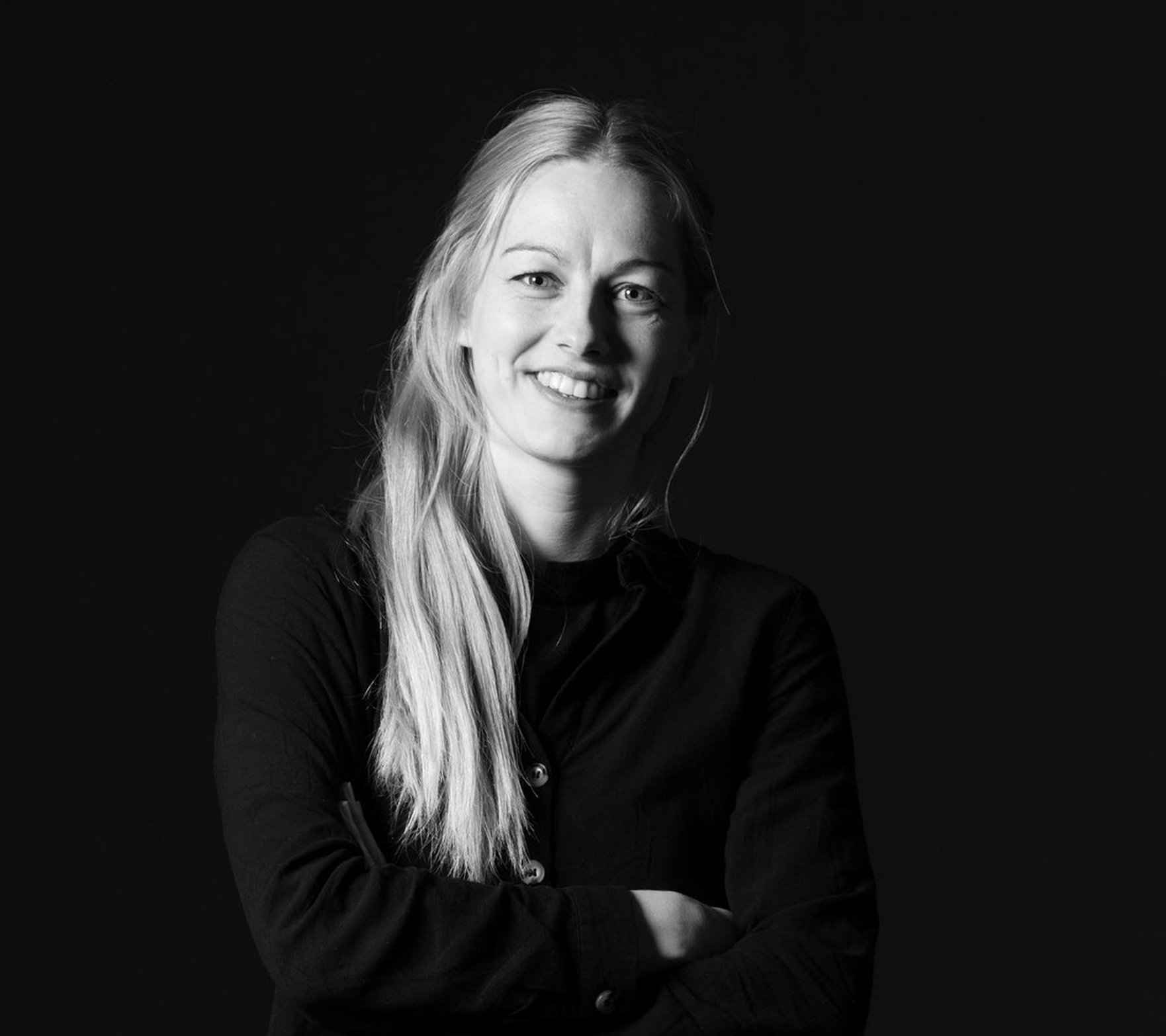 Svartvit porträttbild av en leende kvinna med långt ljust hår och armarna i kors mot en mörk bakgrund.