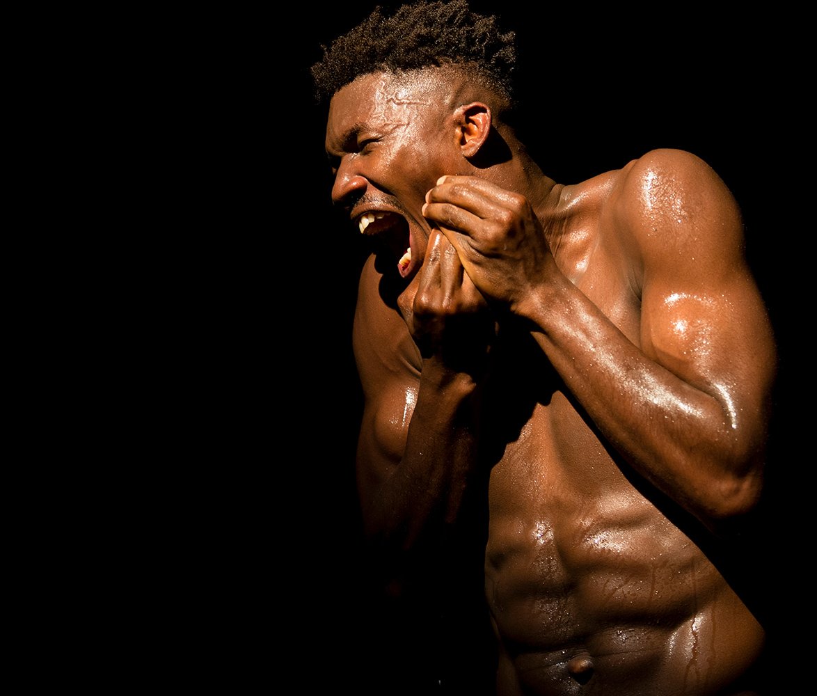 En svettig, barbröstad mörkhyad man med muskulös kropp uttrycker stark känsla genom ansikte och händer mot helt svart bakgrund.