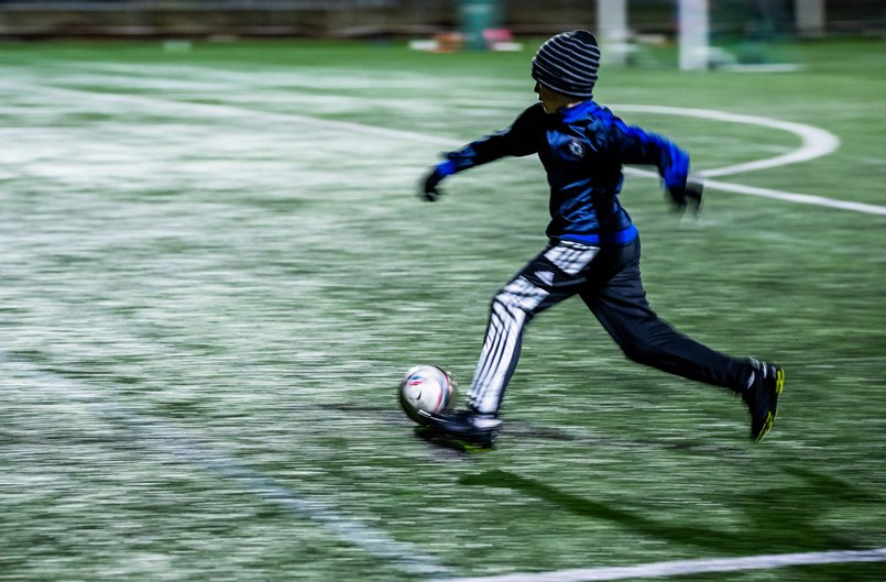 Pojke i mössa och vantar springer med en fotboll i i svagt elljussken.