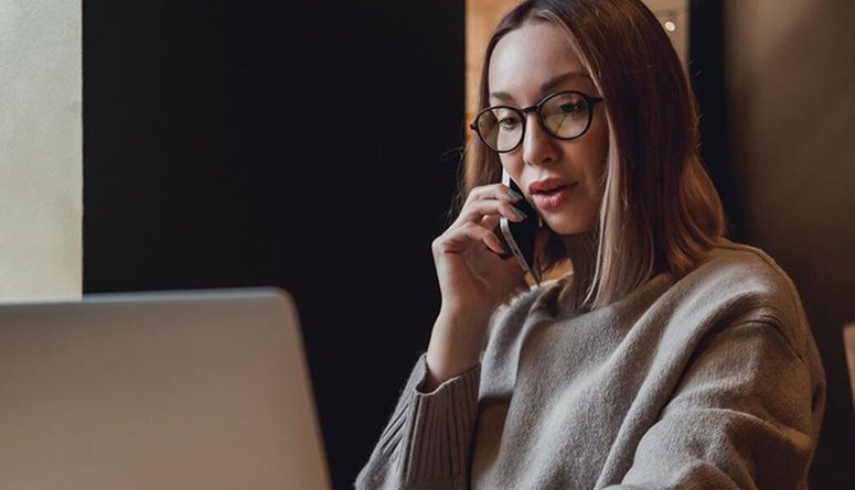 Nainen puhuu puhelimessa samalla kun käyttää tietokonetta.