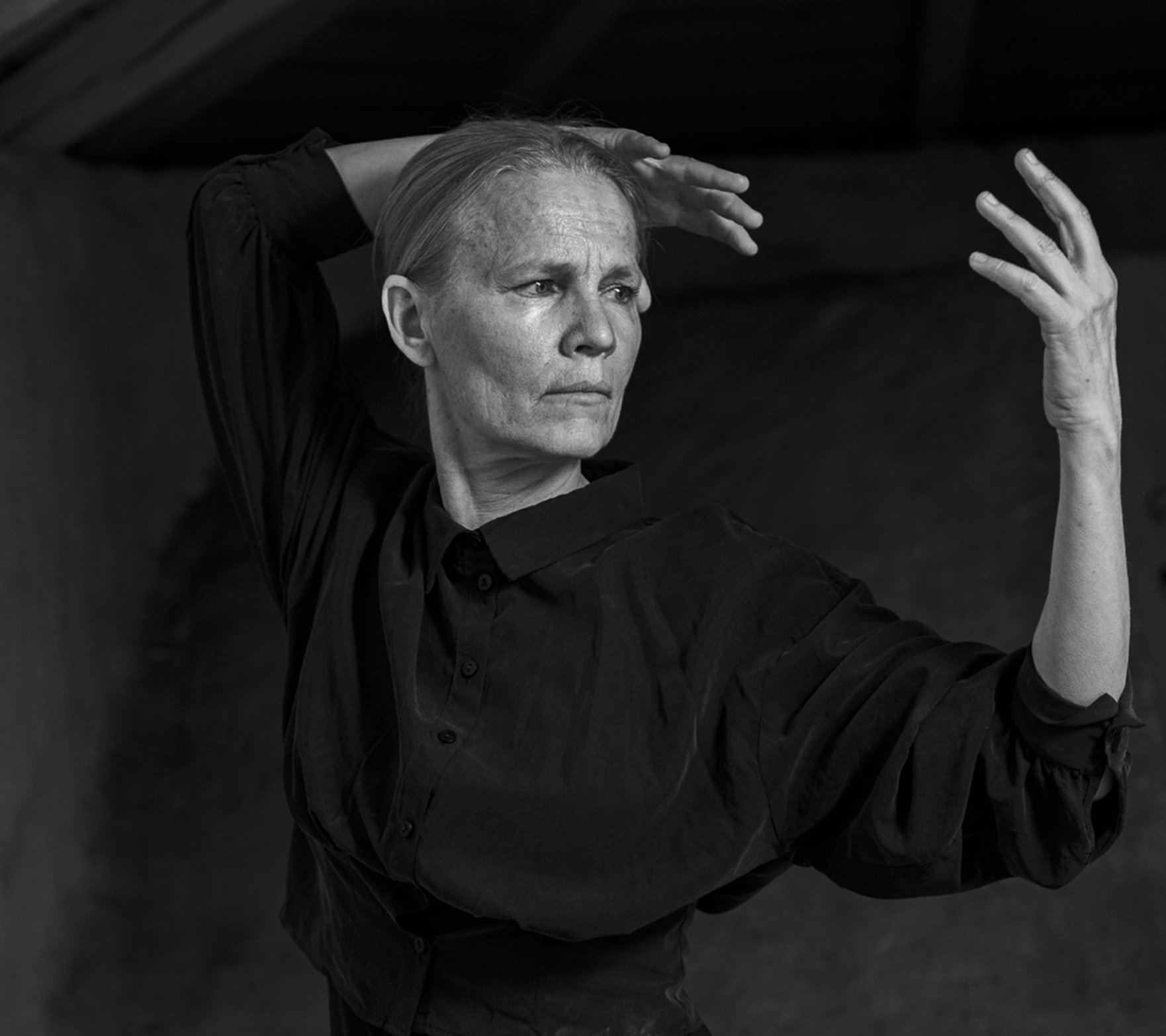 Svartvit bild av en kvinna med håret kammat bakåt, hon har en långärmad skjorta som håller armarna lyfta i en kontrollerad, uttrycksfull danspose inomhus.