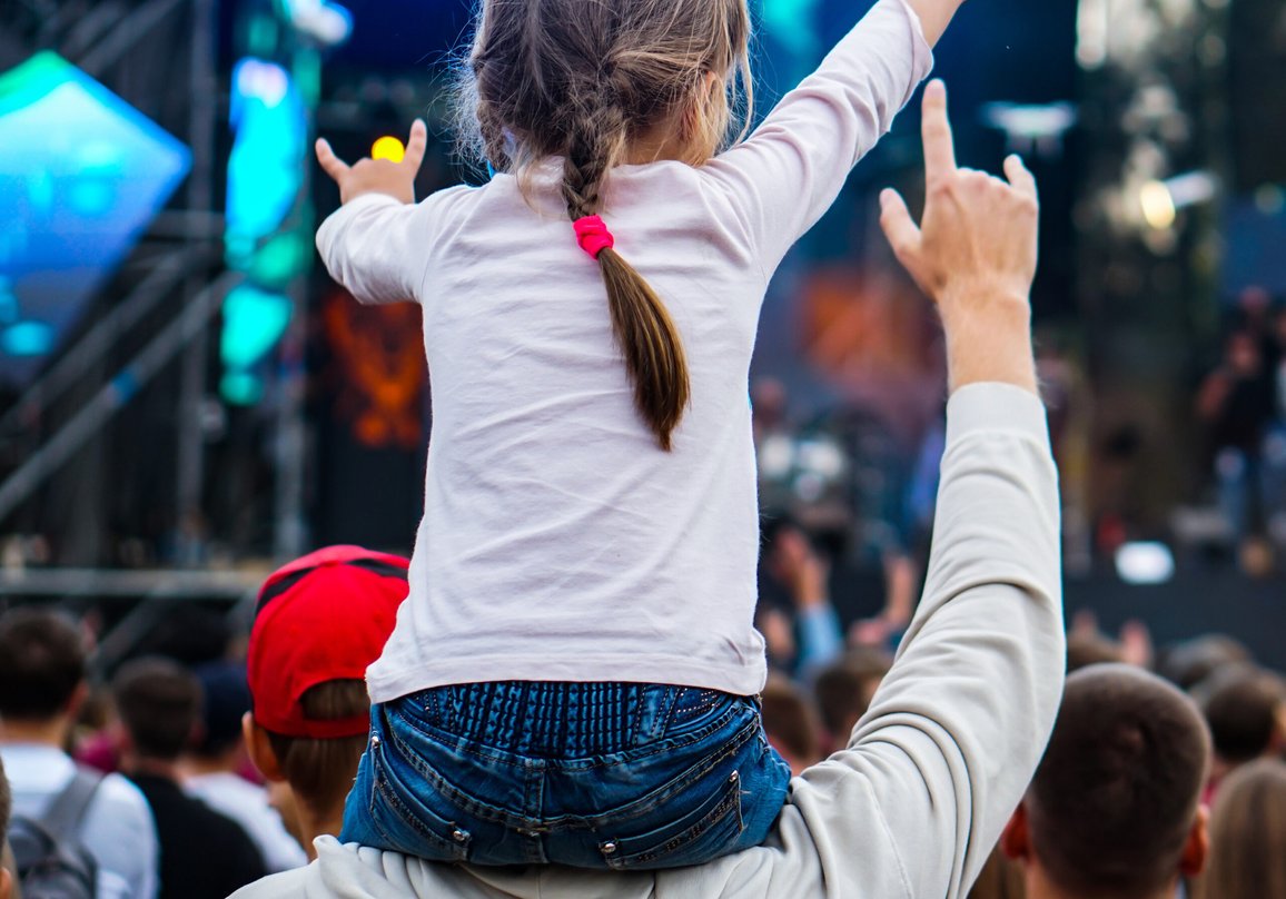 Liten flicka sitter på vuxens man axlar och diggar musik på utomhuskonsert.