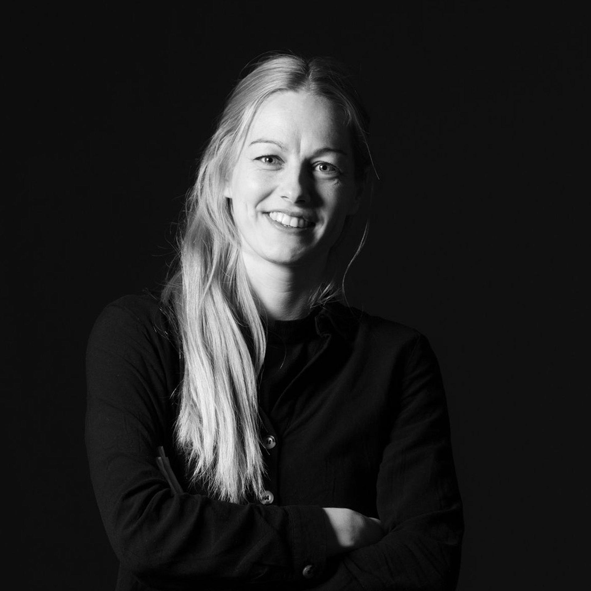 Svartvit porträttbild av en leende kvinna med långt ljust hår och armarna i kors mot en mörk bakgrund.