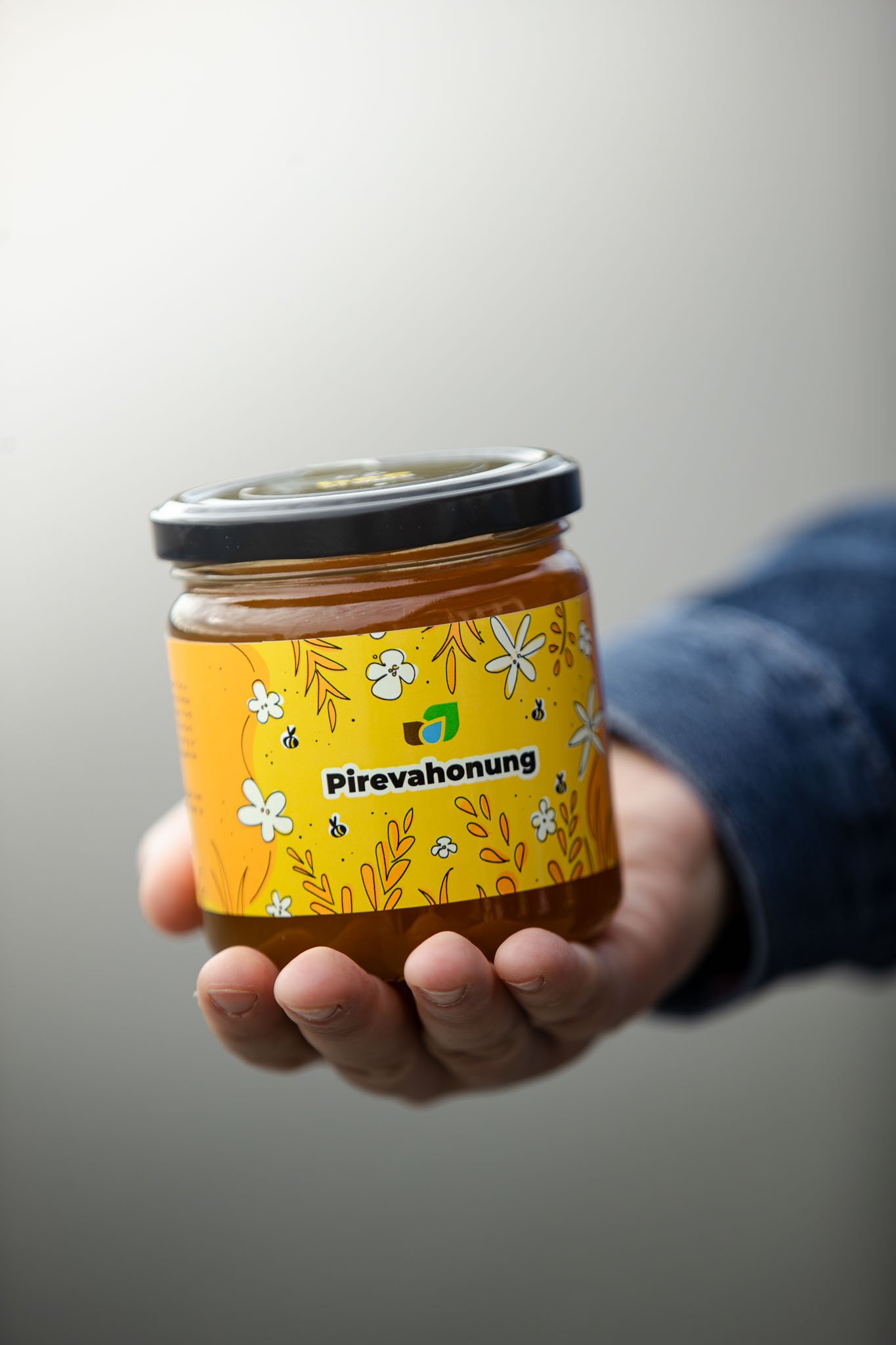 Hand h&aring;ller gul burk med honung m&auml;rkt "Pirevahonung" mot gr&aring; bakgrund.